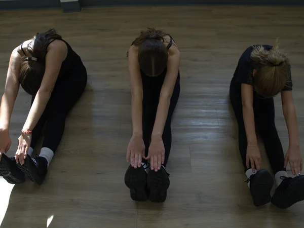 Three young dancers in kids dance classes practicing flexibility by reaching for their toes in a seated position.