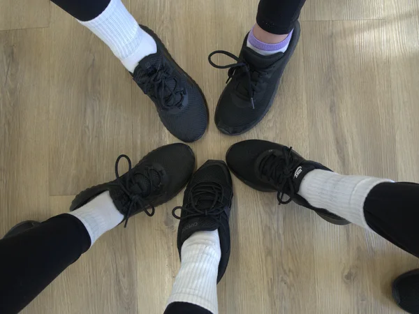 A group of students in kids dance classes standing in a circle showing their black dance sneakers on a wooden floor.