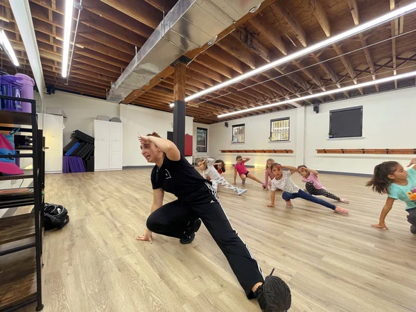 An instructor leading a group of young students through a synchronized leg stretch in kids dance classes.